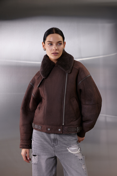 A woman models a thick brown sheepskin aviator bomber with shearling lining, designed in a cropped, oversized silhouette for both warmth and stylish winter layering.