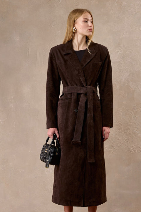 A woman wears an impeccably tailored long chocolate suede coat with sculpted wide shoulders and a cinched waist belt, creating a refined and commanding silhouette.