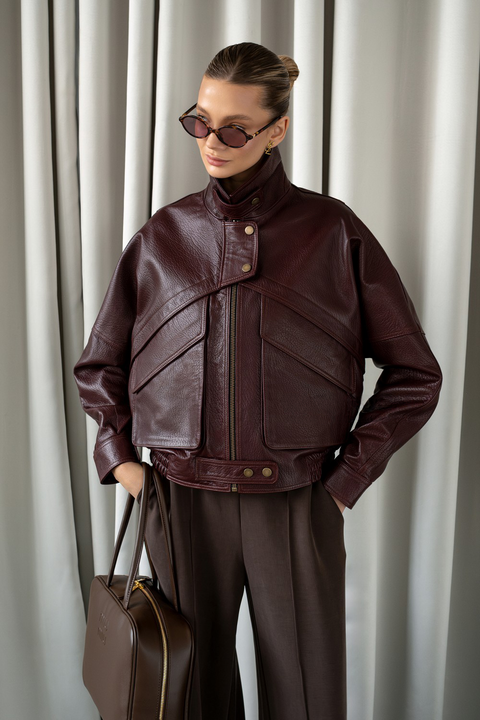 Woman in structured burgundy leather jacket styled with wide-leg trousers and sunglasses, showcasing bold luxury outerwear.