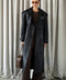 Elegantly dressed, a woman models a black leather trench coat with a classic double-breasted design, sophisticated notched lapels, and belted cuffs made from premium soft leather.